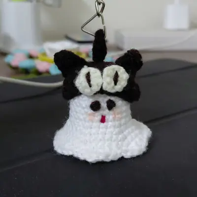 Small white crochet ghost keychain wearing a black cat-ear hood with big eyes